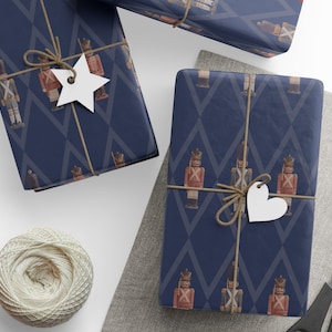 May include: Two rectangular gifts wrapped in dark blue paper featuring a repeating pattern of red and brown nutcracker figures and gray diamond outlines. Each package is tied with brown twine and adorned with a white star or heart tag.