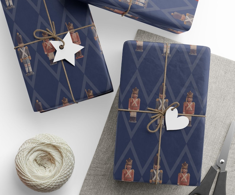 May include: Two rectangular gifts wrapped in dark blue paper featuring a repeating pattern of red and brown nutcracker figures and gray diamond outlines. Each package is tied with brown twine and adorned with a white star or heart tag.