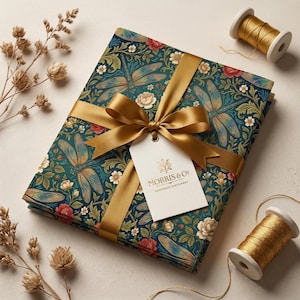 May include: A rectangular stack of stationery wrapped in dark teal paper featuring a repeating pattern of dragonflies, red roses, and white flowers. A gold satin ribbon is tied in a bow around the stack, holding a white tag labeled MORRIS & CO.