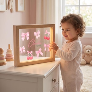 Pode incluir: Uma moldura de madeira quadrada iluminada com um painel de acr&iacute;lico transparente decorado com la&ccedil;os cor-de-rosa, ilustra&ccedil;&otilde;es de cerejas e o nome Claudia Alves em letra cursiva rosa. A moldura tem acabamento em madeira clara e luz LED.