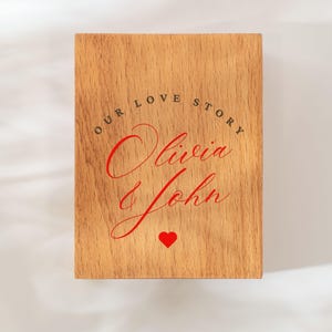 May include: A wooden plaque with the words "OUR LOVE STORY" arched across the top in black, with the names "Olivia John" in red script below. A red heart is at the bottom. The plaque has a light brown wood grain.