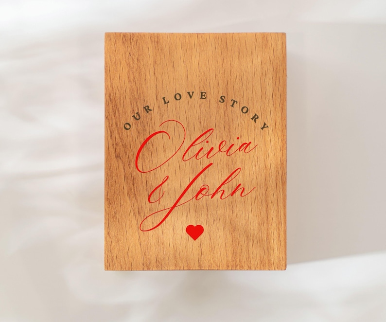 May include: A wooden plaque with the words "OUR LOVE STORY" arched across the top in black, with the names "Olivia John" in red script below. A red heart is at the bottom. The plaque has a light brown wood grain.