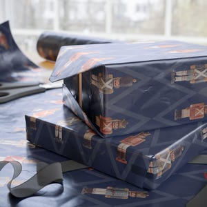May include: Two rectangular boxes wrapped in dark blue paper featuring a repeating pattern of nutcracker figures and light blue diamond outlines. The wrapping paper is also spread out underneath with a pair of scissors and a black ribbon nearby.