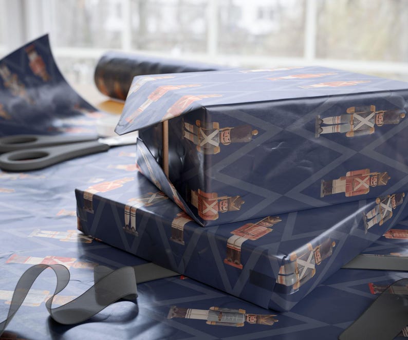 May include: Two rectangular boxes wrapped in dark blue paper featuring a repeating pattern of nutcracker figures and light blue diamond outlines. The wrapping paper is also spread out underneath with a pair of scissors and a black ribbon nearby.