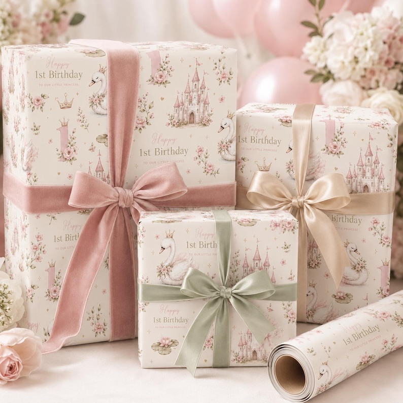 May include: Three gift boxes and a roll of wrapping paper featuring a cream background with a repeating pattern of pink castles, swans wearing crowns, floral arrangements, and the text Happy 1st Birthday, tied with pink, beige, and sage ribbons.