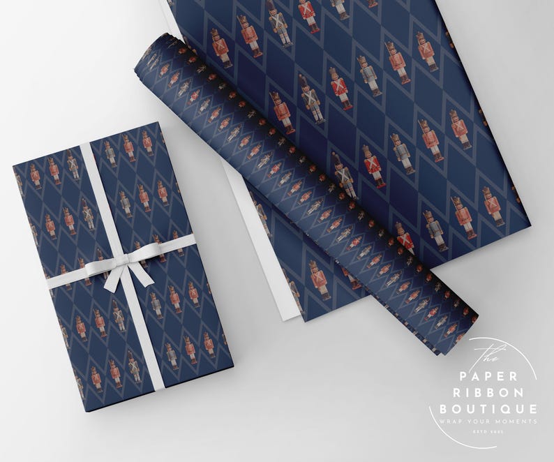 May include: Dark blue wrapping paper featuring a repeating diamond pattern with small nutcracker figures in red and white uniforms. One rectangular gift is wrapped in the paper and tied with a white ribbon. The text The Paper Ribbon Boutique is visible.