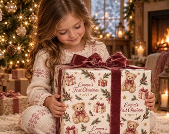 May include: A square gift box wrapped in cream-colored paper featuring a repeating pattern of teddy bears with pink bows, gift boxes, and pine branches. The text reads Emma's First Christmas 2027. A dark pink velvet ribbon is tied in a large bow.