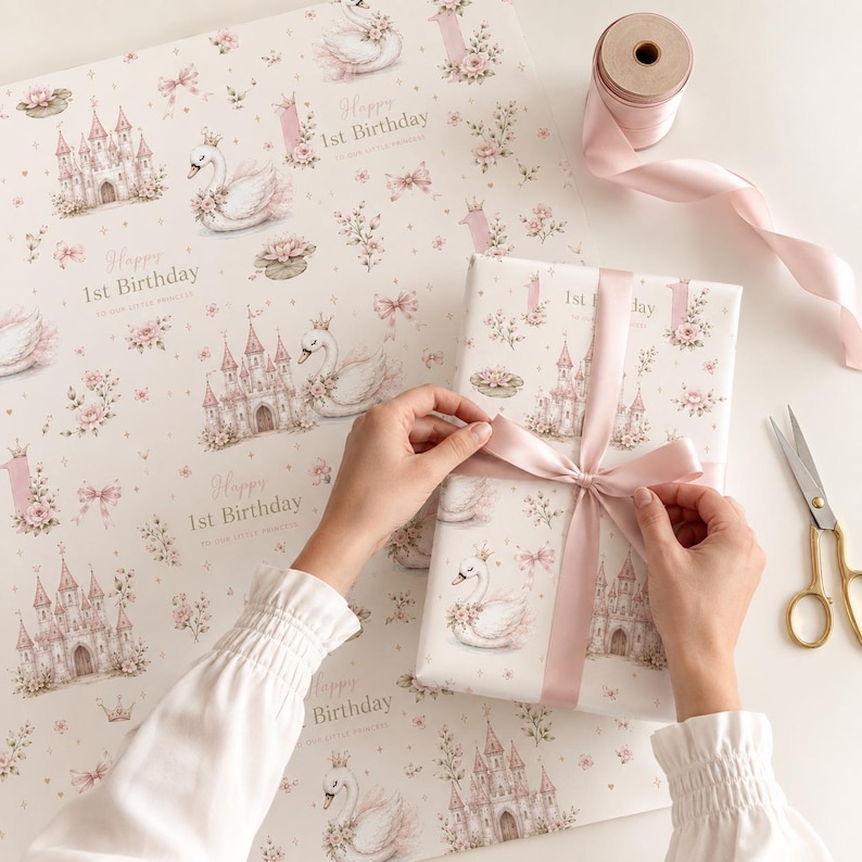 May include: Wrapping paper featuring a repeating pattern of white swans wearing crowns, pink castles, and floral accents with the text Happy 1st Birthday. A gift box wrapped in the same paper is tied with a light pink satin ribbon and a spool of ribbon.