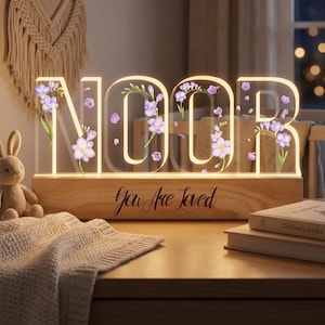 Könnte beinhalten: Ein dekoratives Leuchtschild mit der Aufschrift "NOOR" aus klarem Acryl, verziert mit lila floralen Akzenten. Das Schild steht auf einem Holzsockel mit der Aufschrift "You Are Loved". Ein sanftes Leuchten geht von den Buchstaben aus.