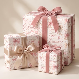 May include: Three gift boxes wrapped in white paper featuring a repeating pattern of pink bows, gold stars, and the text Eleanor and Happy 1st Birthday. The largest and smallest boxes are tied with dusty pink velvet ribbons, while the medium box has a satin ribbon.