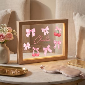 Pode incluir: Uma moldura retangular iluminada com borda de madeira, apresentando la&ccedil;os cor-de-rosa, cerejas e o nome Claudia Alves impresso no vidro. A moldura &eacute; iluminada por dentro, exibindo um design decorativo em uma superf&iacute;cie transparente.