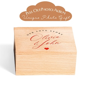 May include: A wooden photo album box with the words "Our Love Story Olivia & John" in red script, and a small red heart. The box is light-colored wood and has a pull-out design. Above the box is a brown cloud-shaped graphic with the text "Pull Out Photo Album Unique Photo Gift."