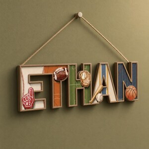 Op de afbeelding: Een decoratieve wandhanger met de naam ETHAN in driedimensionale blokletters, opgehangen aan een touw. Elke letter is versierd met sportthema's, waaronder een football, een honkbalhandschoen, een honkbal, een knuppel en een basketbal.