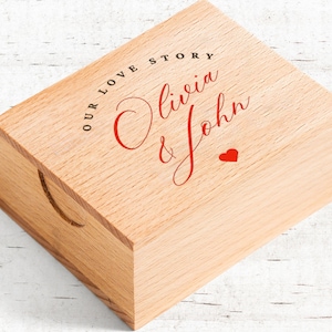 May include: A light-colored wooden box with a recessed handle on the side. The top of the box features the text "OUR LOVE STORY" in black, with the names "Olivia & John" in red script, and a small red heart.