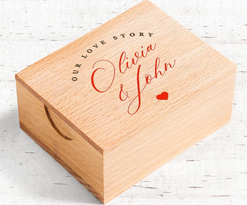 May include: A light-colored wooden box with a recessed handle on the side. The top of the box features the text "OUR LOVE STORY" in black, with the names "Olivia & John" in red script, and a small red heart.