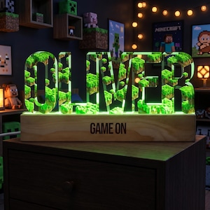 Puede incluir: Un letrero luminoso verde con el nombre "OLIVER" en estilo Minecraft. Las letras están rellenas de bloques verdes pixelados y se asientan sobre una base de madera con la inscripción "GAME ON". El letrero está sobre una superficie de madera oscura.