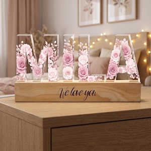 Pode incluir: Um letreiro de acrílico transparente que soletra o nome MILA em letras de bloco, cada uma preenchida com um delicado padrão de rosas cor-de-rosa e flores. As letras estão montadas numa base retangular de madeira clara com o texto gravado We love you.