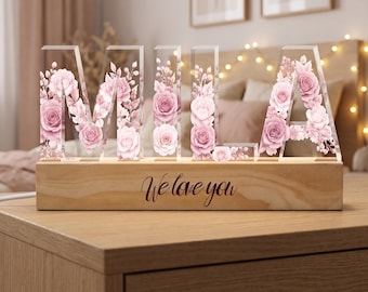 Pode incluir: Um letreiro de acrílico transparente que soletra o nome MILA em letras de bloco, cada uma preenchida com um delicado padrão de rosas cor-de-rosa e flores. As letras estão montadas numa base retangular de madeira clara com o texto gravado We love you.