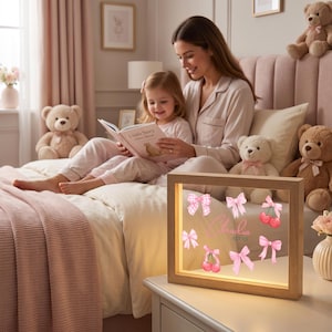Pode incluir: Uma caixa de luz retangular com moldura de madeira e interior iluminado. A frente de vidro exibe o nome Claudia Alves em letra cursiva rosa, rodeado por ilustra&ccedil;&otilde;es de la&ccedil;os cor-de-rosa, fitas xadrez e pares de cerejas vermelhas.