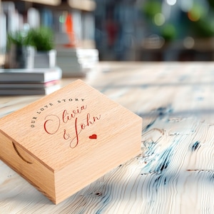 May include: A light-colored wooden box with a lid, featuring the text "OUR LOVE STORY" in a circular font, with "Olivia & John" in cursive and a small red heart. The box has a curved handle cutout on the side.