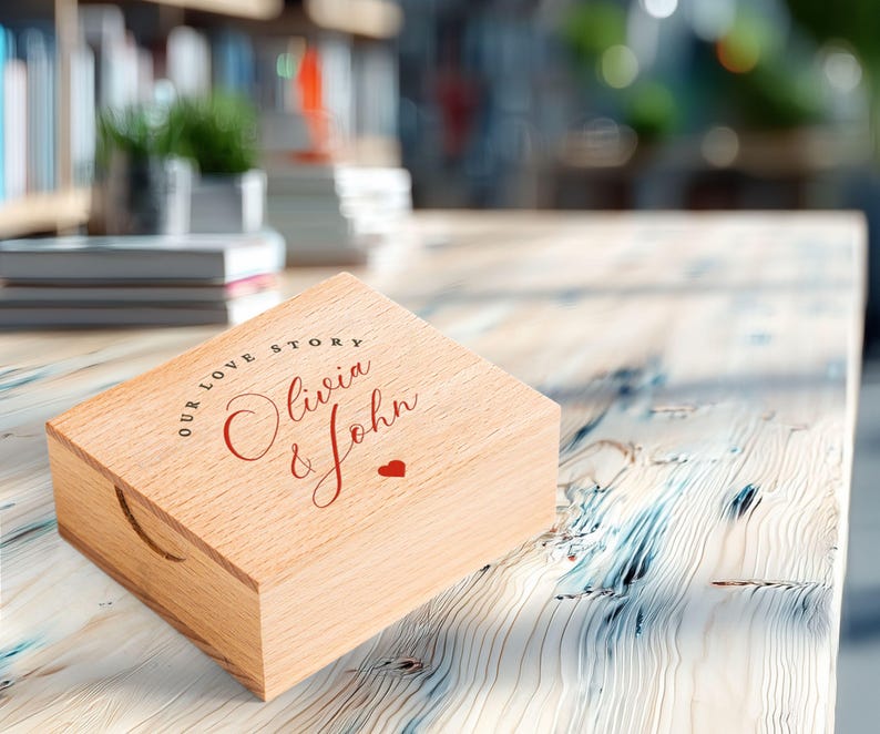 May include: A light-colored wooden box with a lid, featuring the text "OUR LOVE STORY" in a circular font, with "Olivia & John" in cursive and a small red heart. The box has a curved handle cutout on the side.