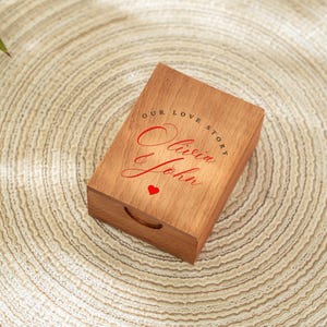 May include: A rectangular wooden box with a handle, featuring the text "OUR LOVE STORY" and the names "Olivia & John" in red script, along with a small red heart. The box is made of light brown wood.