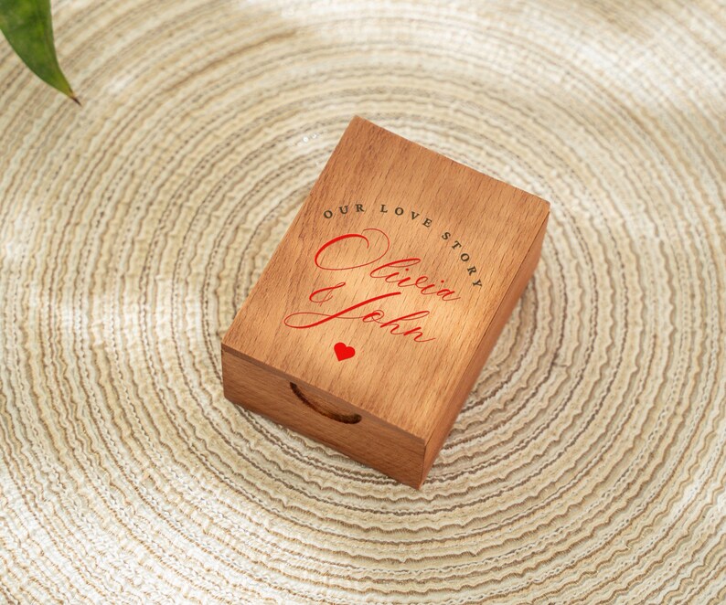 May include: A rectangular wooden box with a handle, featuring the text "OUR LOVE STORY" and the names "Olivia & John" in red script, along with a small red heart. The box is made of light brown wood.