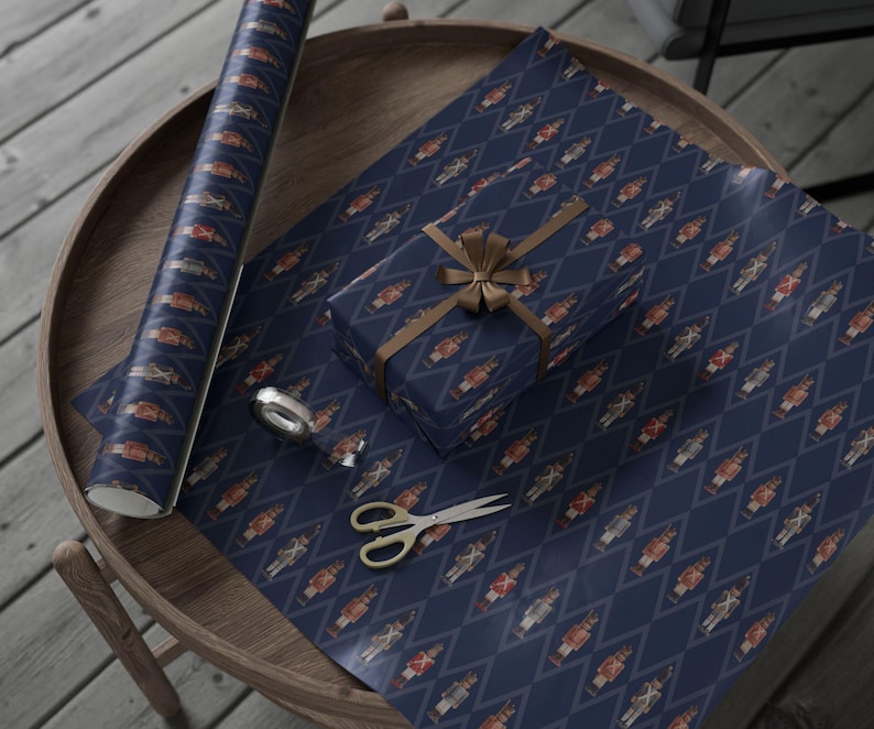 May include: A roll of navy blue wrapping paper featuring a repeating diamond pattern with small nutcracker figures, a gift box wrapped in the same paper with a brown ribbon, a small roll of clear tape, and silver metal scissors.