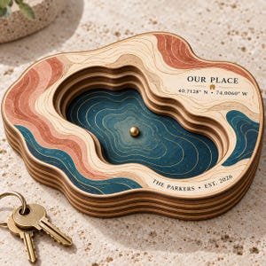 Op de afbeelding: Een gelaagd topografisch houten dienblad met een centraal blauw bekken, een kleine gouden bol en gegraveerde tekst OUR PLACE 40.7128 N 74.0060 W en THE PARKERS EST. 2026. Het dienblad heeft golvende contouren in tinten beige, terracotta en donkerblauw.