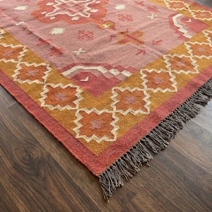 May include: A woven rug with a geometric pattern in shades of pink, orange, and cream. The rug has a fringed edge and is displayed on a dark wood floor. The design features repeating floral and geometric shapes.