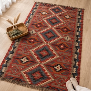 May include: A rectangular area rug featuring a repeating geometric diamond pattern in shades of red, blue, black, and cream. The rug has a fringed edge on the top and bottom and is decorated with a woven texture throughout the fabric surface.
