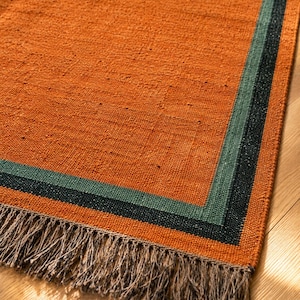 May include: A rectangular woven rug featuring a large burnt orange center panel bordered by a sage green stripe and a dark charcoal black outer stripe. The rug is finished with a textured fringe along the edge in a neutral light brown tone.