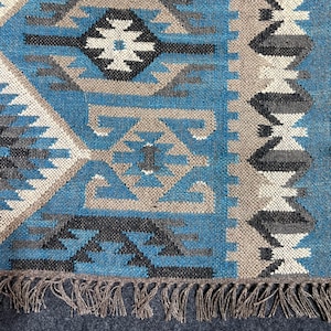 May include: Close-up of a blue, gray, and beige woven rug with a geometric pattern. The rug features diamond and triangle shapes in various shades, with a fringe along the bottom edge. The rug is suitable for home decor.