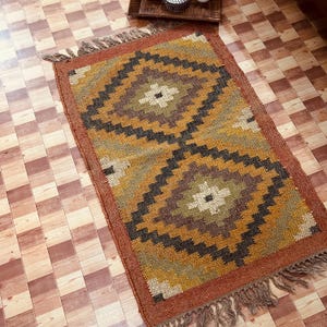 Può includere: Un tappeto rettangolare con un motivo geometrico a rombi nei toni del marrone, oro, nero e verde. Il tappeto ha un bordo con frange e un bordo marrone rossiccio. Realizzato con fibre naturali.