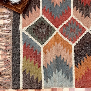 Könnte beinhalten: Ein farbenfroher geometrischer Teppich mit einem Rautenmuster in Rot-, Blau-, Grün-, Orange- und Brauntönen. Der Teppich hat einen Fransenrand und einen dunklen Rand. Das Design zeigt ineinandergreifende Formen und einen neutralen weißen Rand.