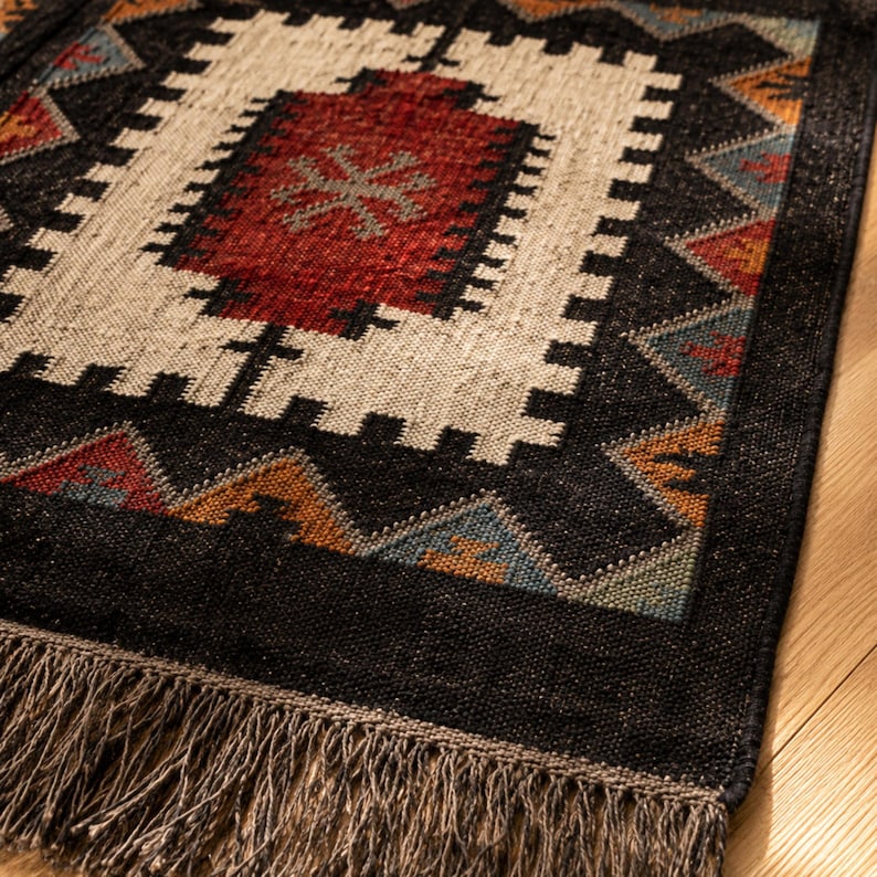 Close up of handwoven black wool jute rug showing geometric tribal pattern in rust burgundy ivory and teal, natural fiber flatweave texture