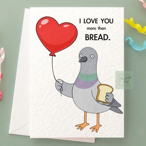 May include: A white greeting card with the text "I LOVE YOU more than BREAD." features a cartoon pigeon holding a red heart-shaped balloon and a slice of bread. The pigeon is gray with purple and green accents.