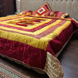 Może przedstawiać: Łóżko przykryte żywym, pikowanym narzutą. Projekt zawiera centralny kwadrat z kwiatowymi wzorami, otoczony żółtymi, bordowymi i wzorzystymi obramowaniami. Na łóżku spoczywa pasująca poduszka z geometrycznym wzorem.