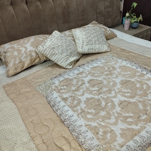 May include: A bed featuring a beige and white floral quilt and matching pillows. The pillows are square and rectangular, with decorative stitching and embellishments. The headboard is a dark brown, padded design.