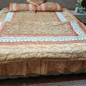 May include: A gold and peach-toned bedspread set. The set includes a patterned comforter, matching pillowcases, and a bed skirt. The fabric has a soft, velvety texture, and the design features floral embroidery.