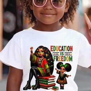 May include: White t-shirt featuring a vibrant graphic with the text "EDUCATION IS FREEDOM." The design includes an illustration of a woman and a child, both with dark hair and wearing colorful outfits. The woman is sitting on a stack of books.