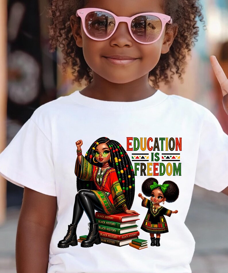 May include: White t-shirt featuring a vibrant graphic with the text "EDUCATION IS FREEDOM." The design includes an illustration of a woman and a child, both with dark hair and wearing colorful outfits. The woman is sitting on a stack of books.