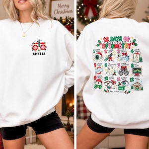 May include: A white crewneck sweatshirt featuring a small graphic of optometry equipment and the name Amelia on the front left chest. The back displays a large, colourful 12 Days of Optometry graphic with twelve numbered illustrations and text.