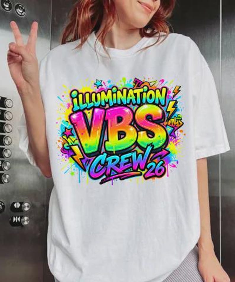 May include: White t-shirt with a colorful graphic design. The design features the words "ILLUMINATION VBS CREW 26" in vibrant rainbow colors, surrounded by stars, lightning bolts, and graffiti-style accents. The shirt is a casual, unisex style.