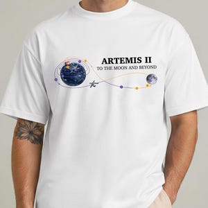 May include: White t-shirt featuring the text "ARTEMIS II TO THE MOON AND BEYOND." The design includes a stylized depiction of Earth, a spacecraft, and the moon, connected by a curved path with purple and gold accents.