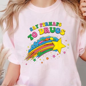 Puede incluir: Camiseta rosa claro con el texto "SAY PERHAPS TO DRUGS" en letras coloridas. Un arcoíris y una estrella amarilla adornan la parte inferior de la camiseta, con pequeñas estrellas dispersas.