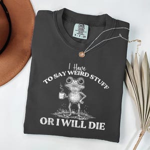 Puede incluir: Camiseta gris oscuro Comfort Colors con el texto "I Have To Say Weird Stuff Or I Will Die" y una rana de dibujos animados sosteniendo una taza de café. Un collar plateado con un colgante y un sombrero marrón también están en la imagen.