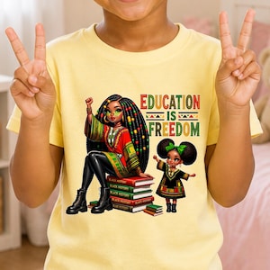 May include: Yellow t-shirt featuring a graphic of two Black girls, one seated on books labeled "Black History." The text "Education is Freedom" is displayed in red, green, and black. The girls are wearing colorful outfits and boots.