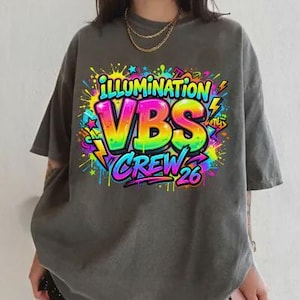 May include: Dark gray t-shirt with a colorful graphic that reads "ILLUMINATION VBS CREW 26". The graphic features rainbow-colored letters and neon accents, with lightning bolts and starbursts. The shirt has short sleeves and a relaxed fit.