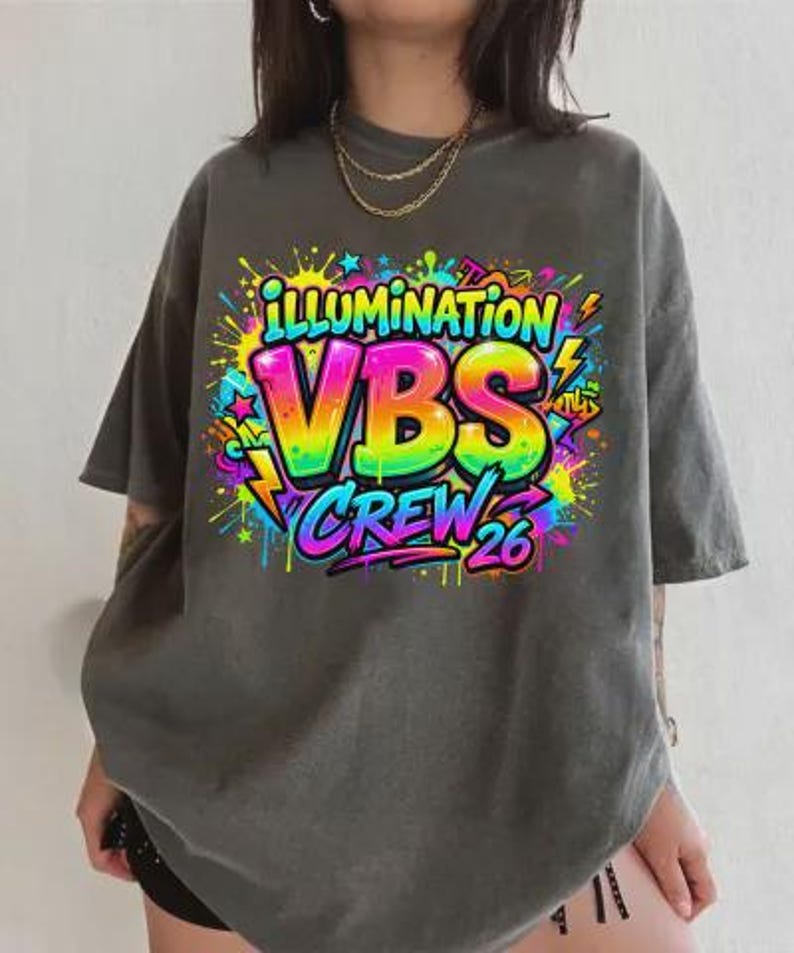May include: Dark gray t-shirt with a colorful graphic that reads "ILLUMINATION VBS CREW 26". The graphic features rainbow-colored letters and neon accents, with lightning bolts and starbursts. The shirt has short sleeves and a relaxed fit.